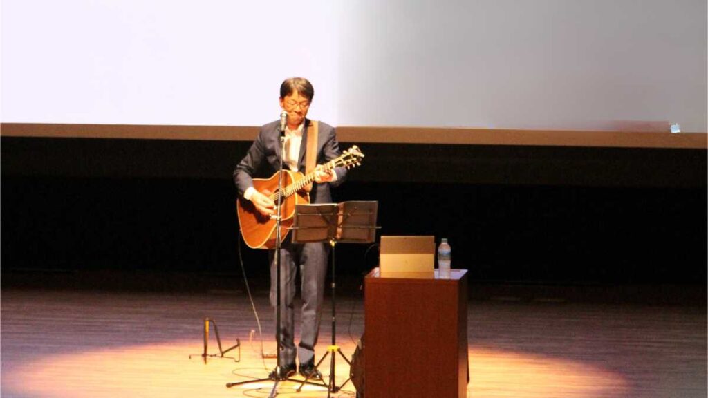 講演でオリジナルソングを歌う石川達之